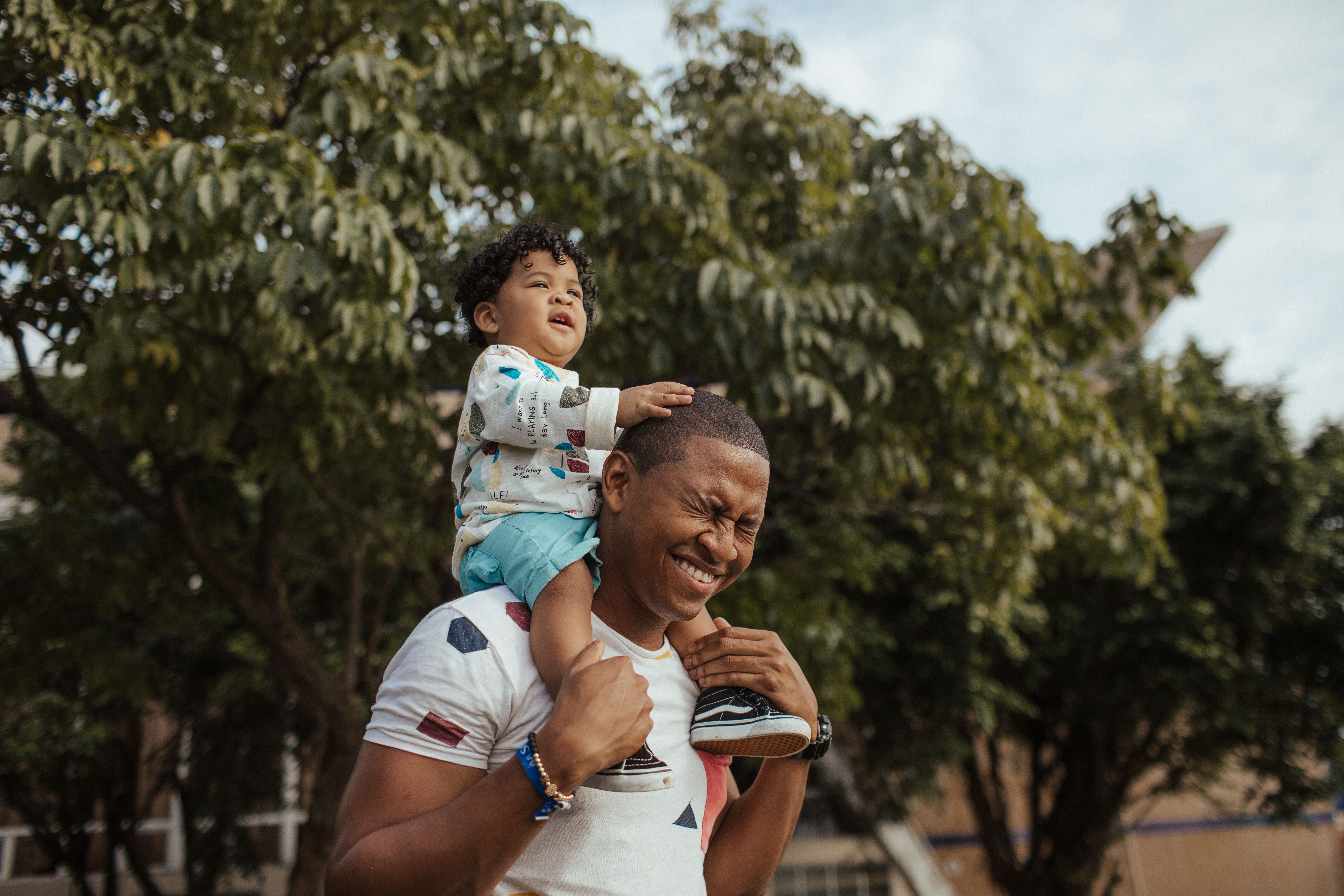 Father outside, in front of trees, laughing with child sitting on his shoulder.
